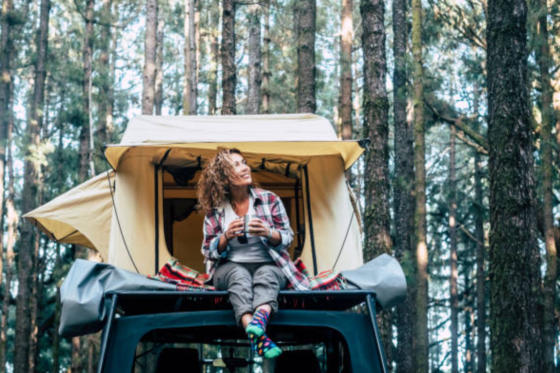 A woman with curly hair sits in front of her tent with a cup of coffee in the morning