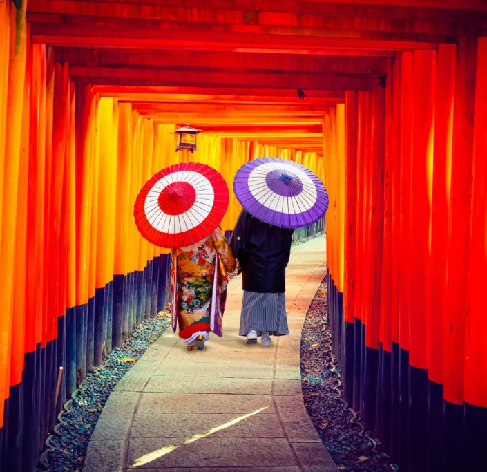 Two people in kimono with umbrellas walking in the Fushimi Inari Shrine gates.